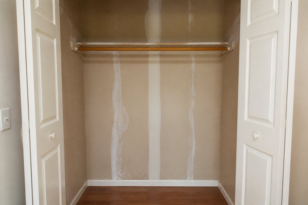 An empty closet with shelving removed, showing bare drywall and a wooden rod, ready for conversion into a home office
