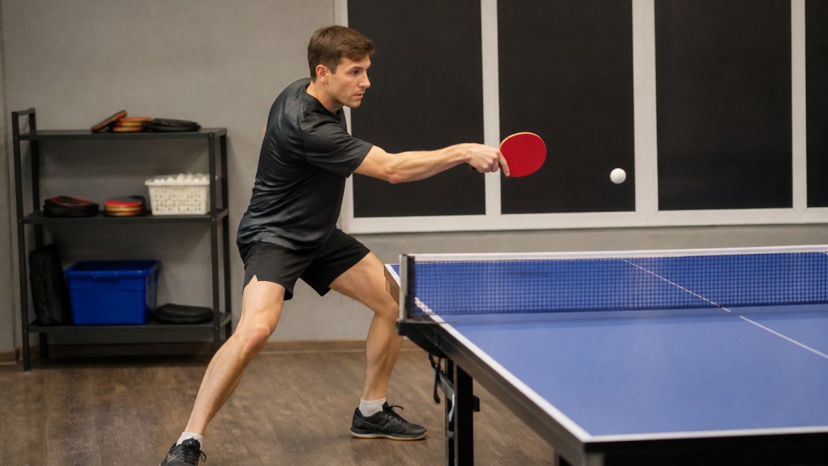 Player in athletic stance mid-forehand stroke at home table tennis table, demonstrating proper footwork and body position during a solo training drill, with organized equipment visible in the background