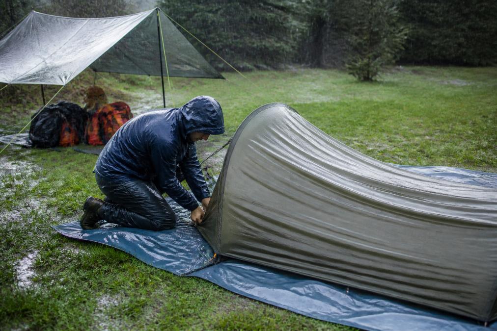 Camper kneeling in wet grass pitching a tent rainfly first during a rainstorm with gear protected nearby.