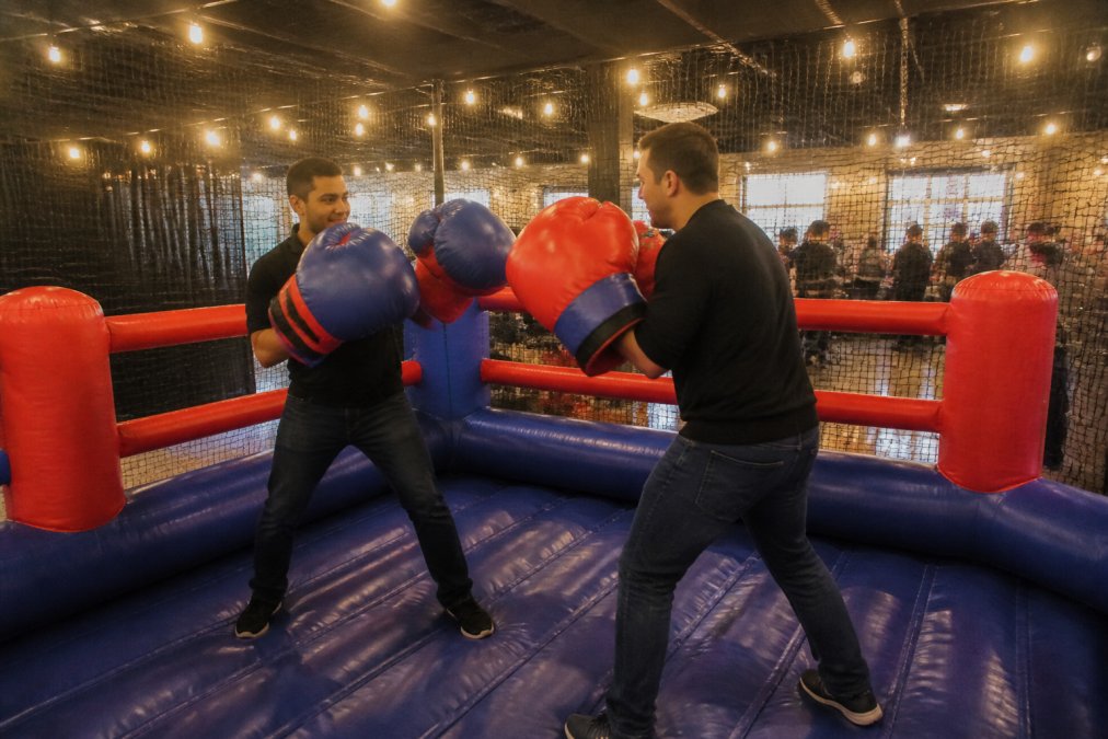 Professional demonstrating inflatable boxing ring technique in modern event space with safety netting visible, showing the oversized gloves and cushioned flooring around the perimeter