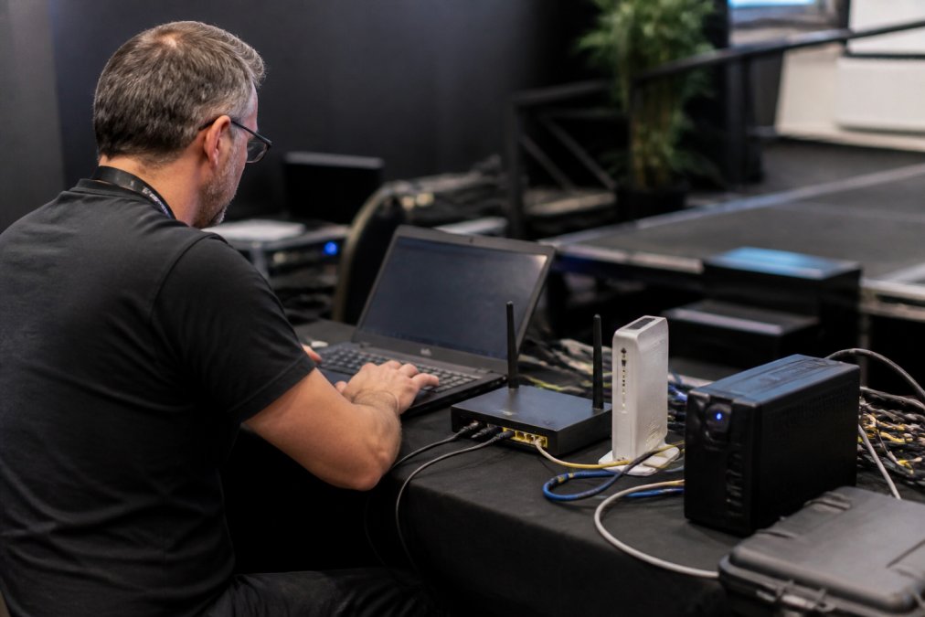 A broadcast technician at a live event venue checking a laptop connected to a mobile broadband router and a secondary modem, with cables and a small UPS unit visible on the table beside them