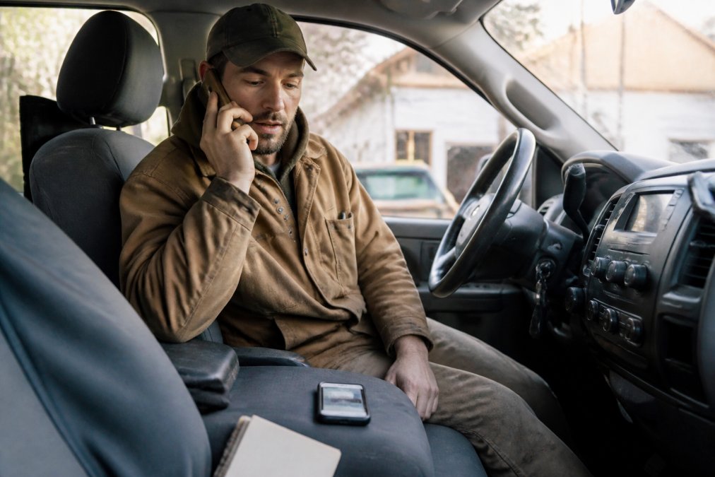 A contractor in a work jacket sitting in the cab of a pickup truck, phone pressed to his ear mid-call, glancing at his phone screen on the seat beside him, conveying urgency, natural daylight through the windshield