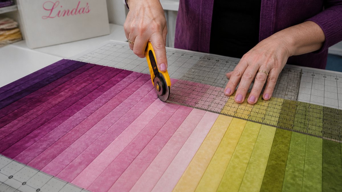 Close-up of hands using a rotary cutter and quilting ruler to cut fabric strips in graduated colors arranged on a self-healing mat