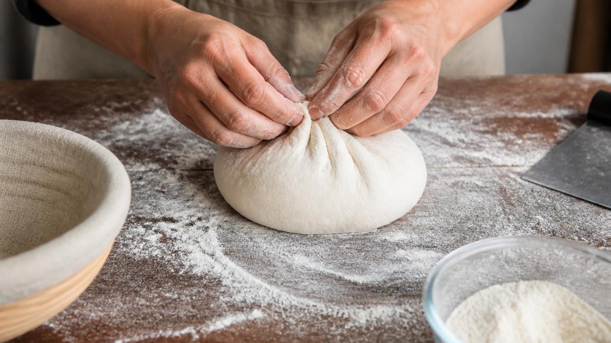 Hands tucking and pulling fermented dough to create surface tension while shaping a round sourdough boule