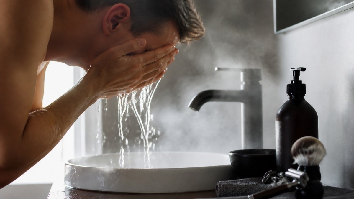 Close-up of a man splashing warm water on his face over a bathroom sink, steam rising, preparing his skin before shaving