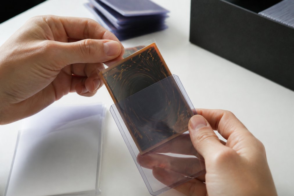 Close-up of hands placing a trading card into a penny sleeve, then sliding it into a top loader, showing proper card protection technique with natural lighting on a white surface