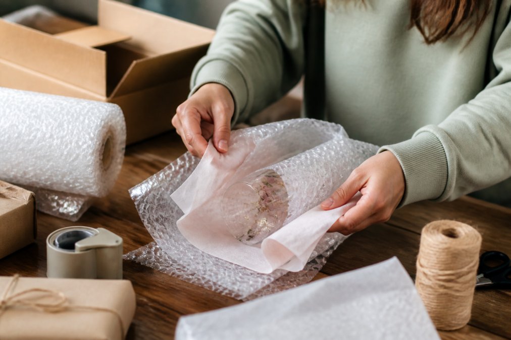 Hands carefully wrapping a delicate personalized necklace in tissue paper, then bubble wrap, on a clean work surface with packing materials organized nearby