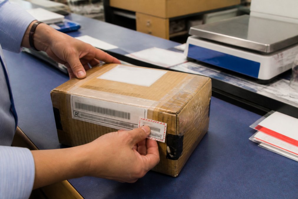 Postal clerk's hands placing a registered mail label and signature confirmation sticker on a carefully packaged gold shipment wrapped in corrugated cardboard with visible filament tape and security seals, with the USPS scale and counter visible in background