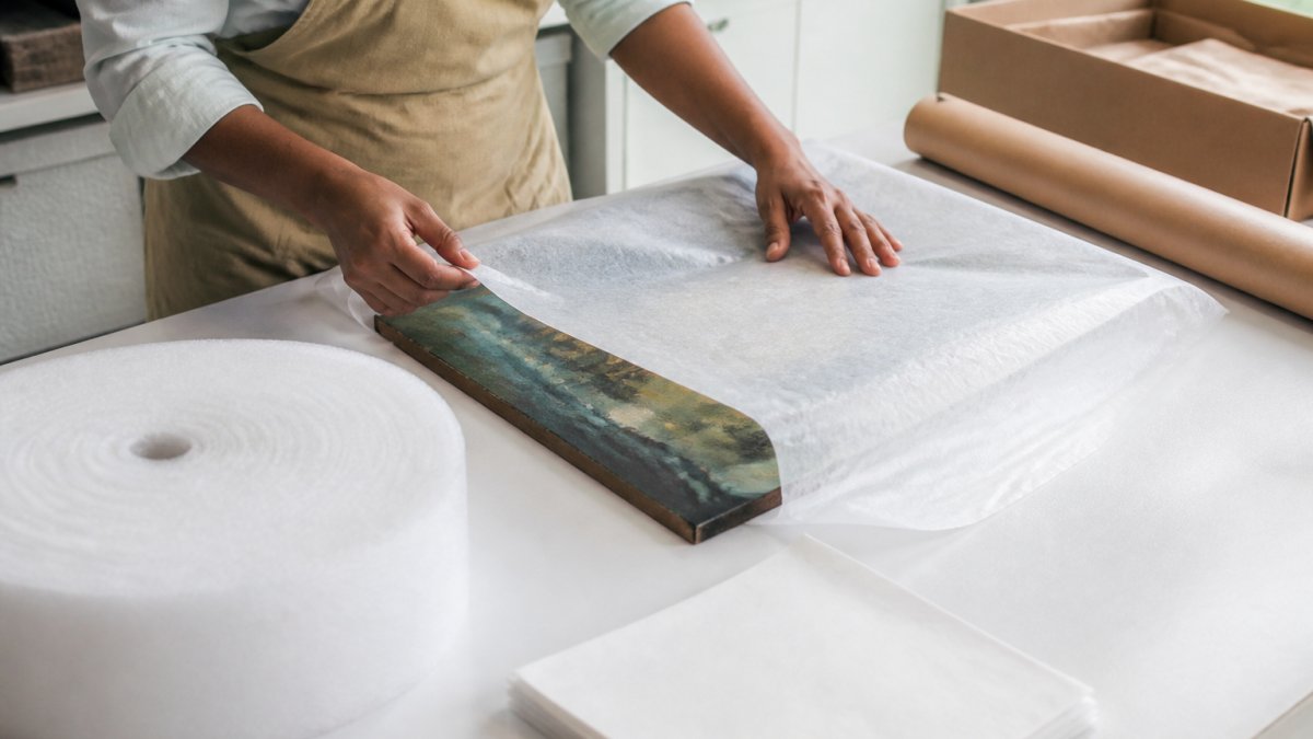 Professional carefully wrapping an original canvas painting with glassine paper and acid-free materials on a clean white work surface, hands shown gently handling the artwork