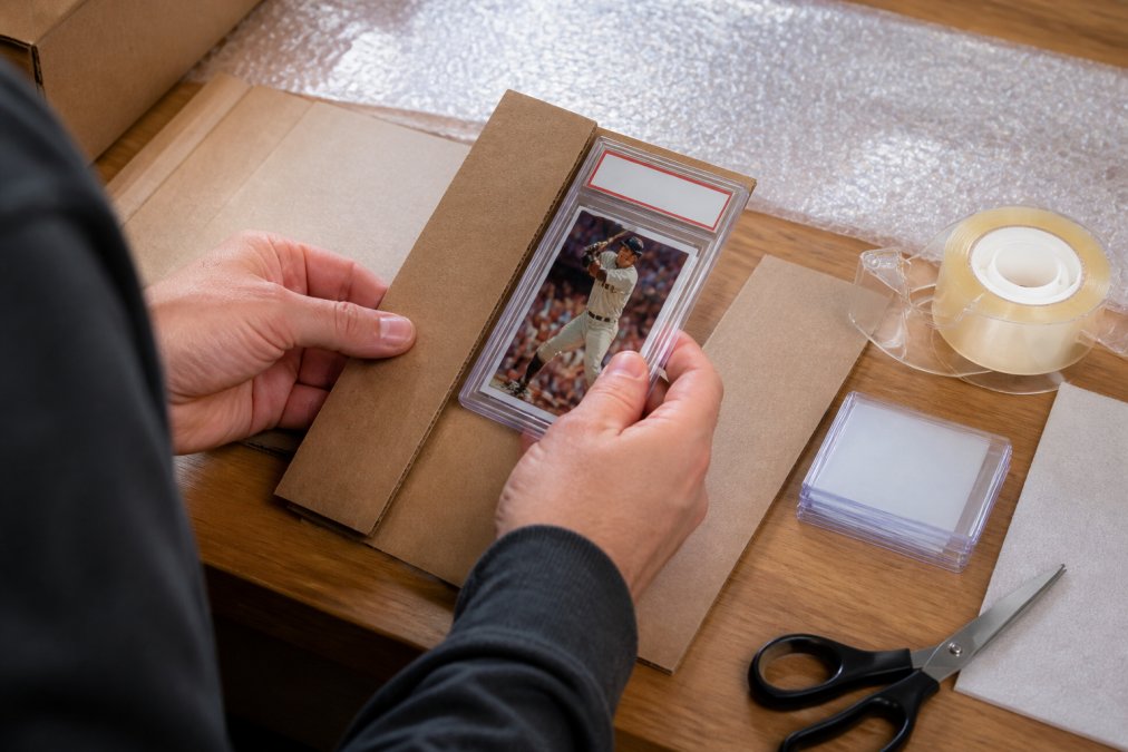 Close-up of hands carefully placing a graded sports card in its protective slab into a rigid cardboard sandwich with bubble wrap visible in the background on a clean work surface