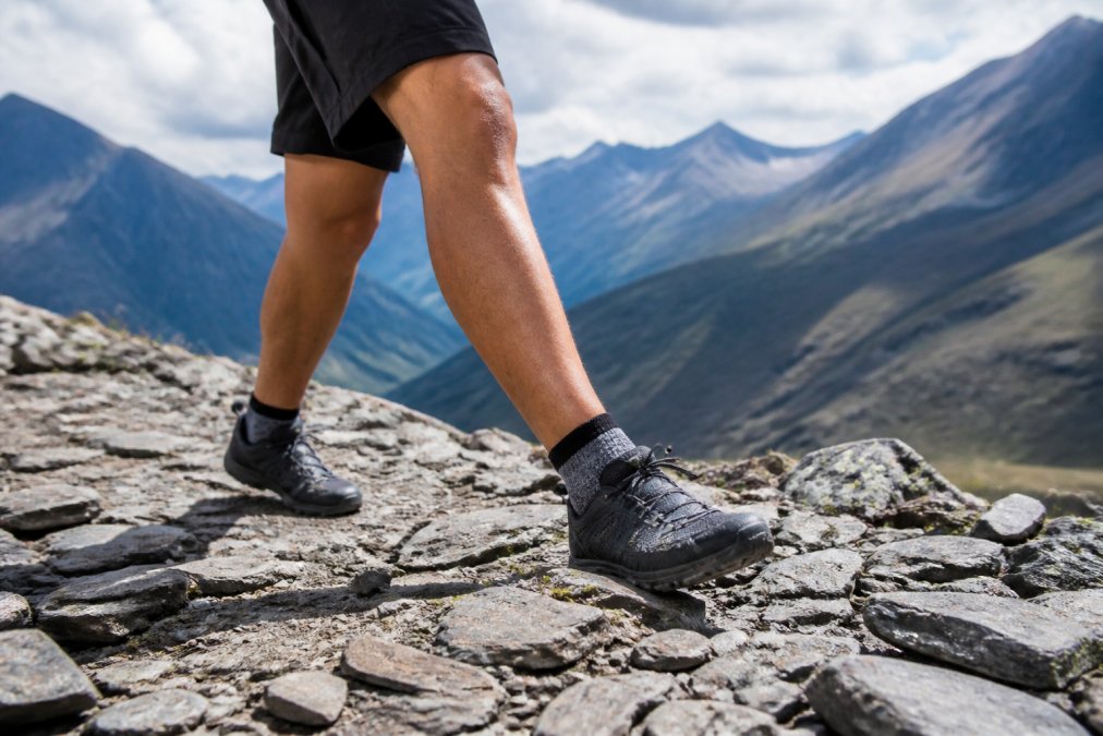 Trail runner wearing RockResist low-cut socks visible on rocky mountain terrain, feet clearly shown on loose gravel and stone, demonstrating durability during active hiking movement with natural outdoor lighting
