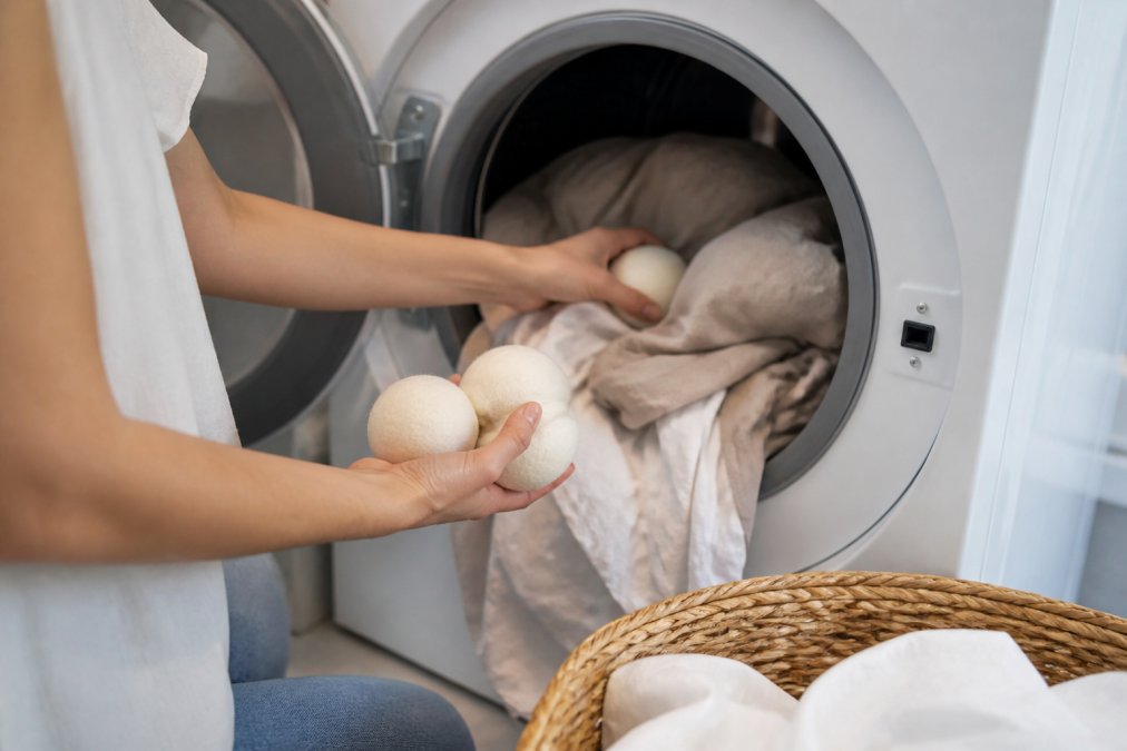 Hands placing wool dryer balls into a dryer filled with linen clothing in a bright laundry room