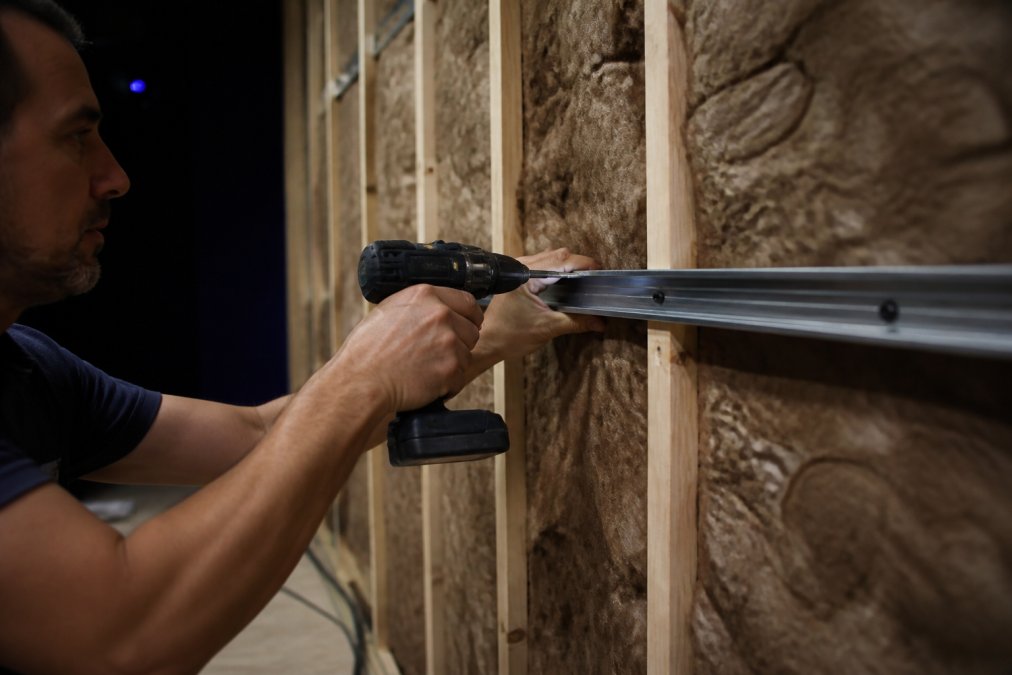 Close-up of a worker's hands installing a metal resilient channel onto wooden wall studs in a dimly lit home theater room under construction, with insulation visible in the cavities