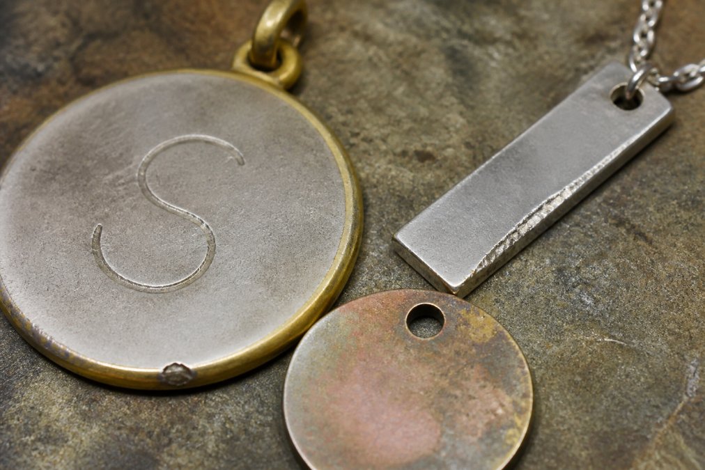 Close-up of personalized jewelry pieces showing shallow engraving, rough metal edges, and visible base metal discoloration under natural light on a jeweler's work surface
