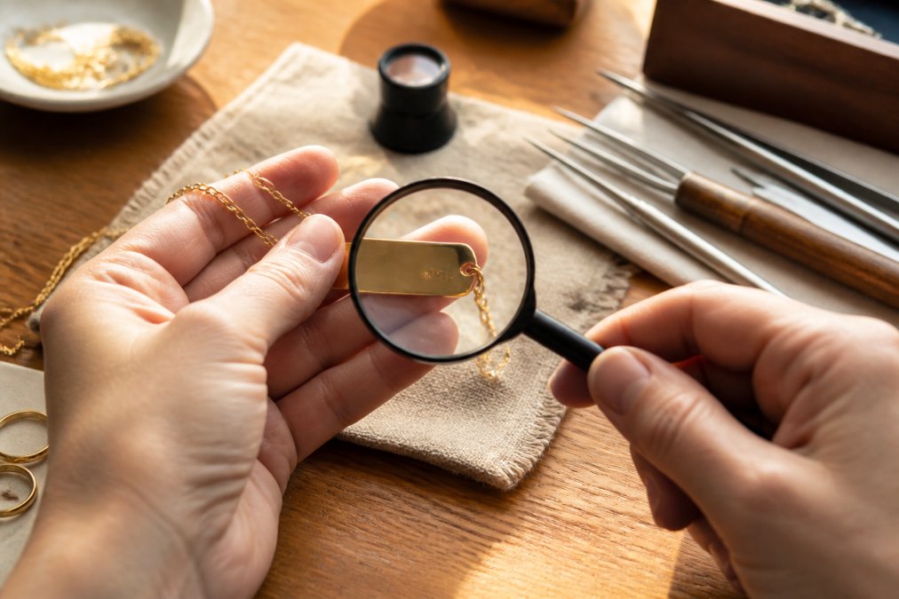 Hands holding a personalized gold necklace next to a magnifying glass, examining hallmarks and maker's marks stamped on the metal surface under bright natural light