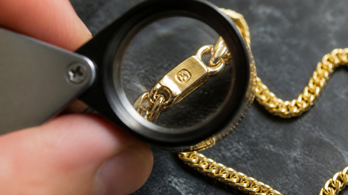 Close-up photograph of a jeweler's loupe held over the clasp of a gold necklace, revealing a small stamped hallmark on the metal tab
