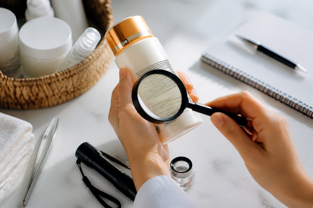 Close-up of hands carefully examining a beauty product bottle under bright natural lighting, checking the batch number label, packaging details, holographic seal, and font clarity with magnifying glass
