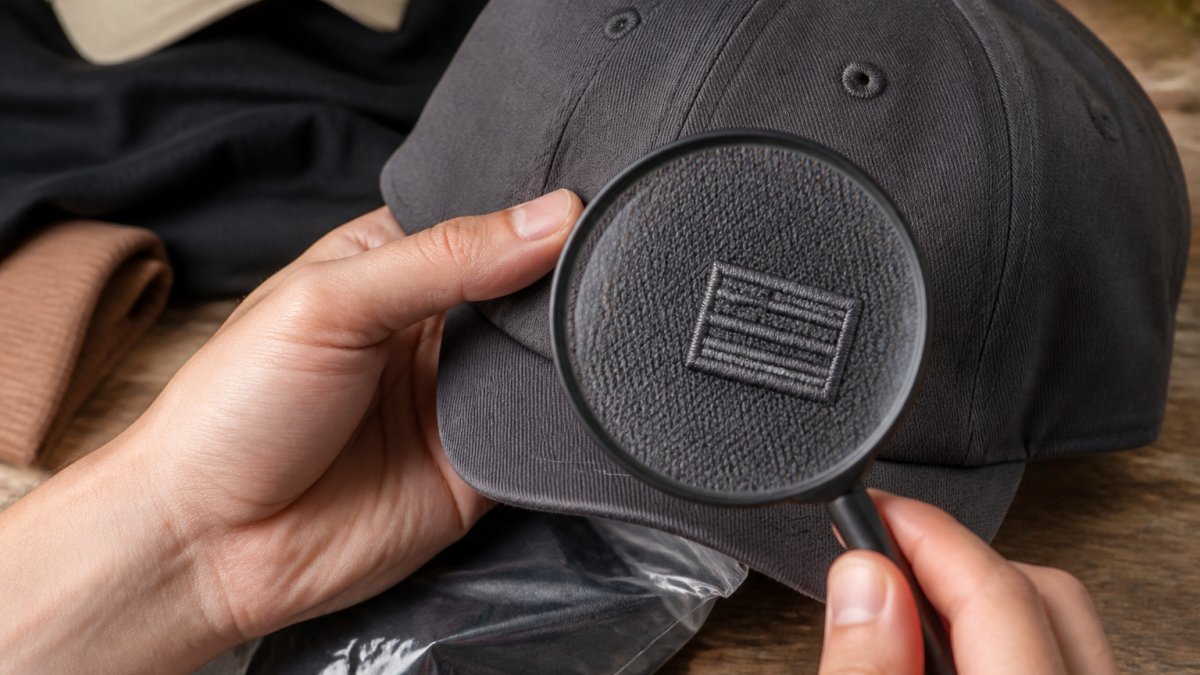 Close-up of hands holding a designer baseball cap, with a magnifying glass inspecting the embroidered logo and stitching on the front panel