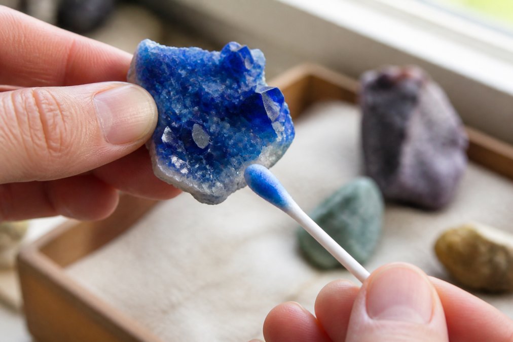 Close-up of a hand holding a vivid blue crystal specimen over a white cotton swab, showing blue color transfer on the swab from the acetone test, bright natural window lighting