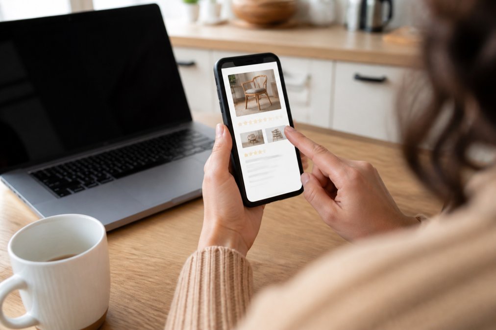 Close-up of a person's hands holding a smartphone displaying a product page with star ratings, sitting at a kitchen table with a laptop nearby