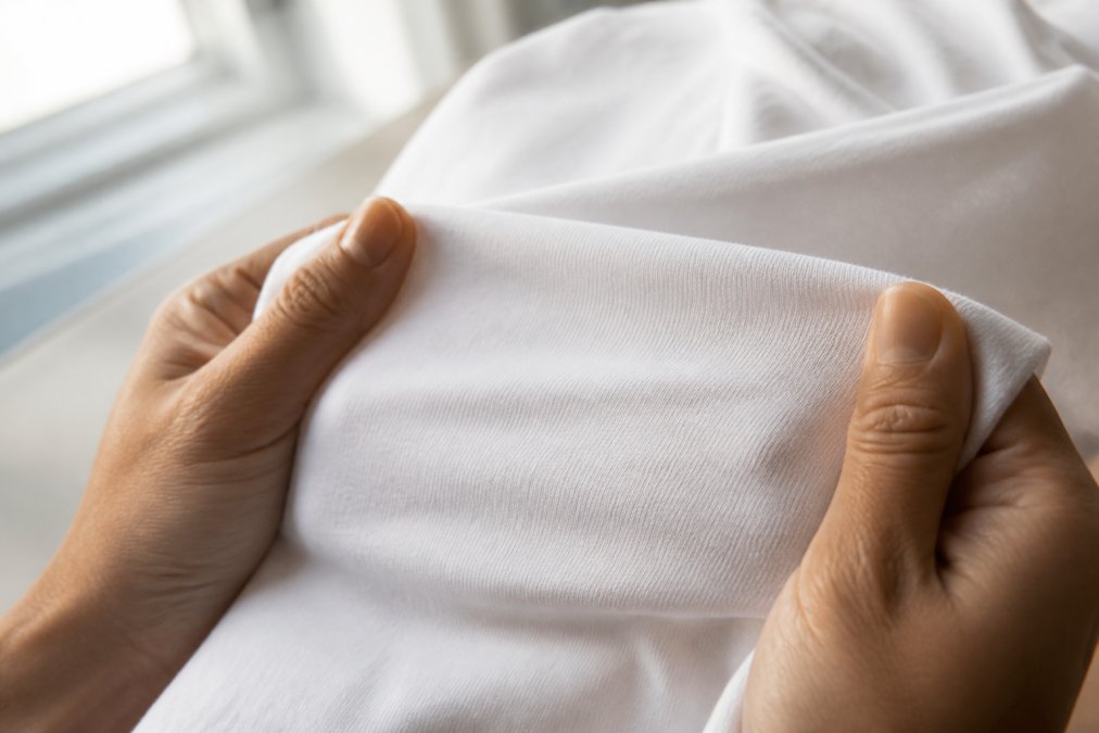 Close-up of two hands gently stretching a white cotton t-shirt fabric between fingers, showing the fine weave texture under soft natural window lighting