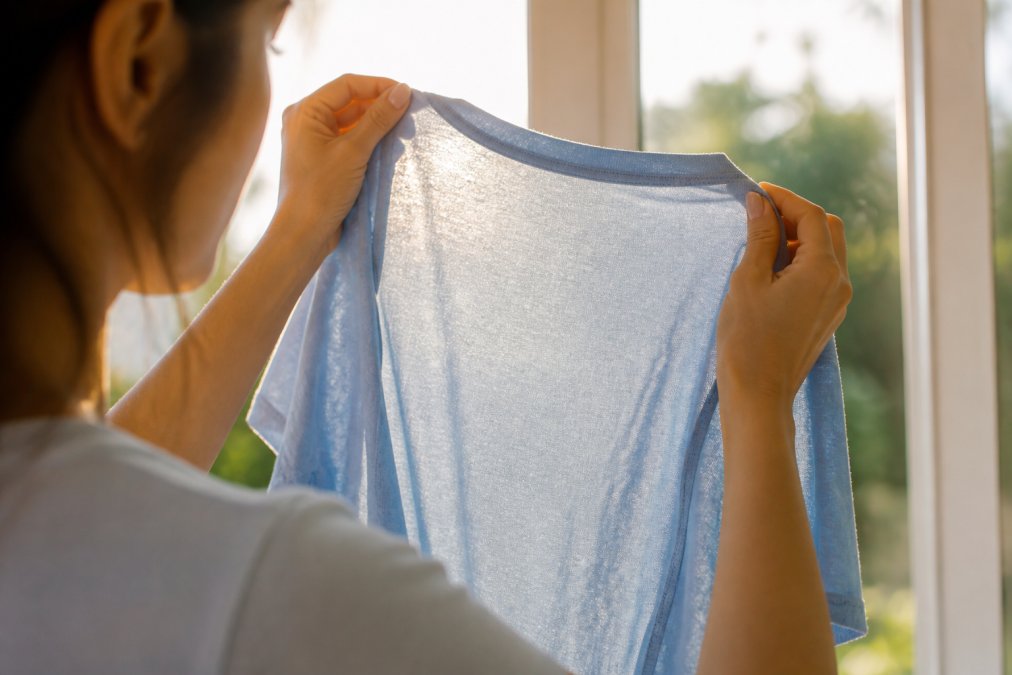Hands holding a light blue t-shirt against a window to inspect the weave density of the high quality fabric.