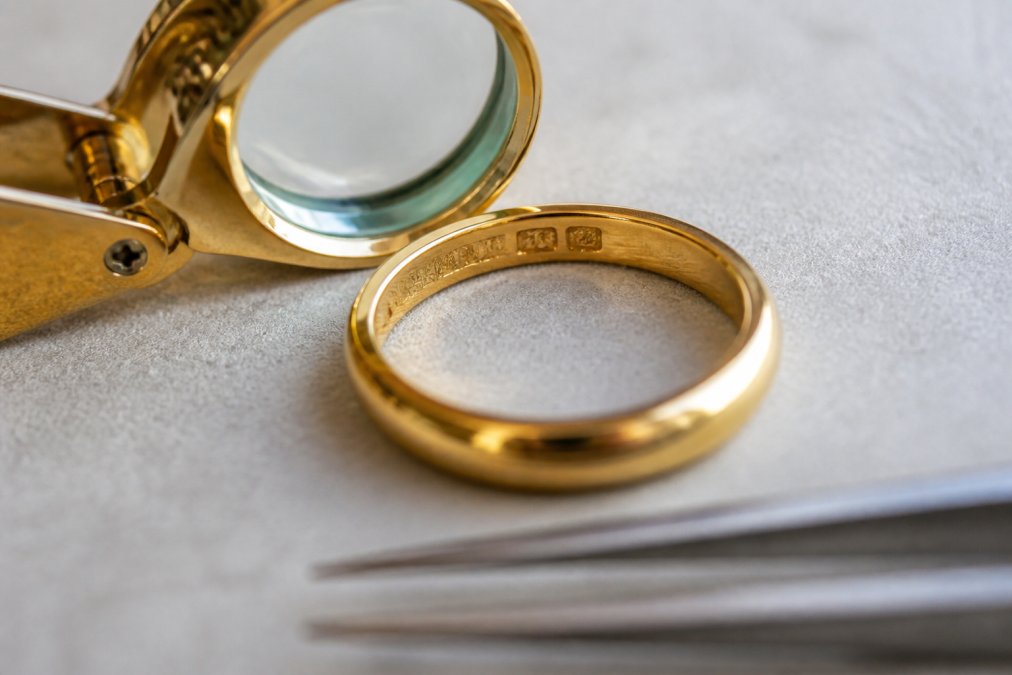 Close-up of gold ring showing hallmark stamps and engravings under bright natural light with magnifying glass nearby