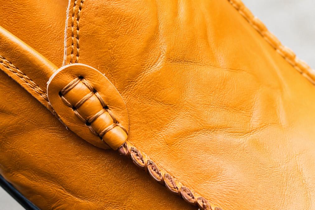 Macro close-up of a full grain leather surface showing natural pores, subtle fat wrinkles, and irregular grain pattern on a tan leather loafer under bright directional lighting