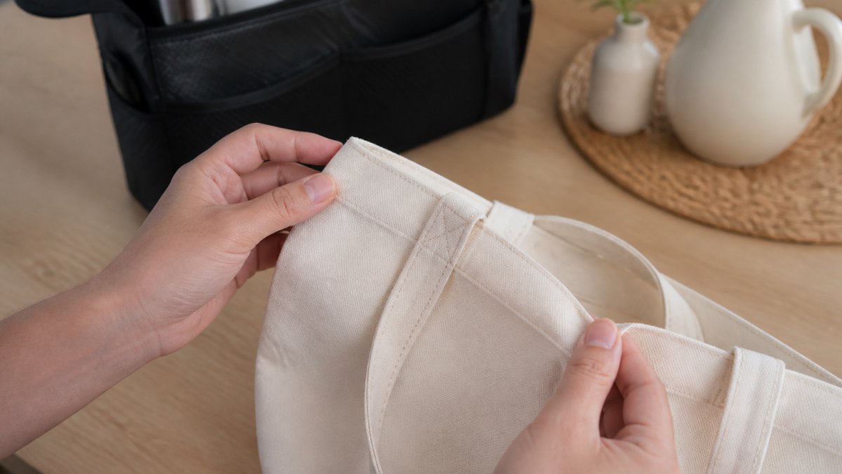 Close-up of hands inspecting the stitching and fabric of a tote bag on a clean wooden table, with a car organizer and decorative home items softly blurred in the background