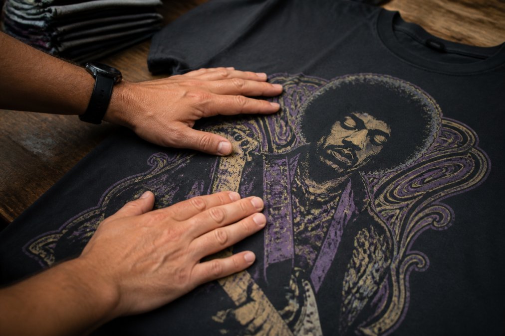Hands examining and feeling the texture of a screen printed design on a vintage-style rock graphic tee, demonstrating tactile assessment of ink thickness and flexibility