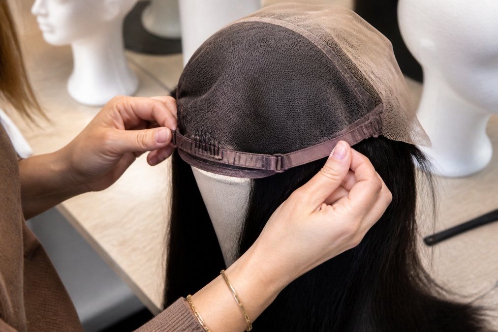 Close-up of a woman's hands examining the inside cap of a lace front wig on a styling block, showing the lace mesh and adjustable straps in soft natural light