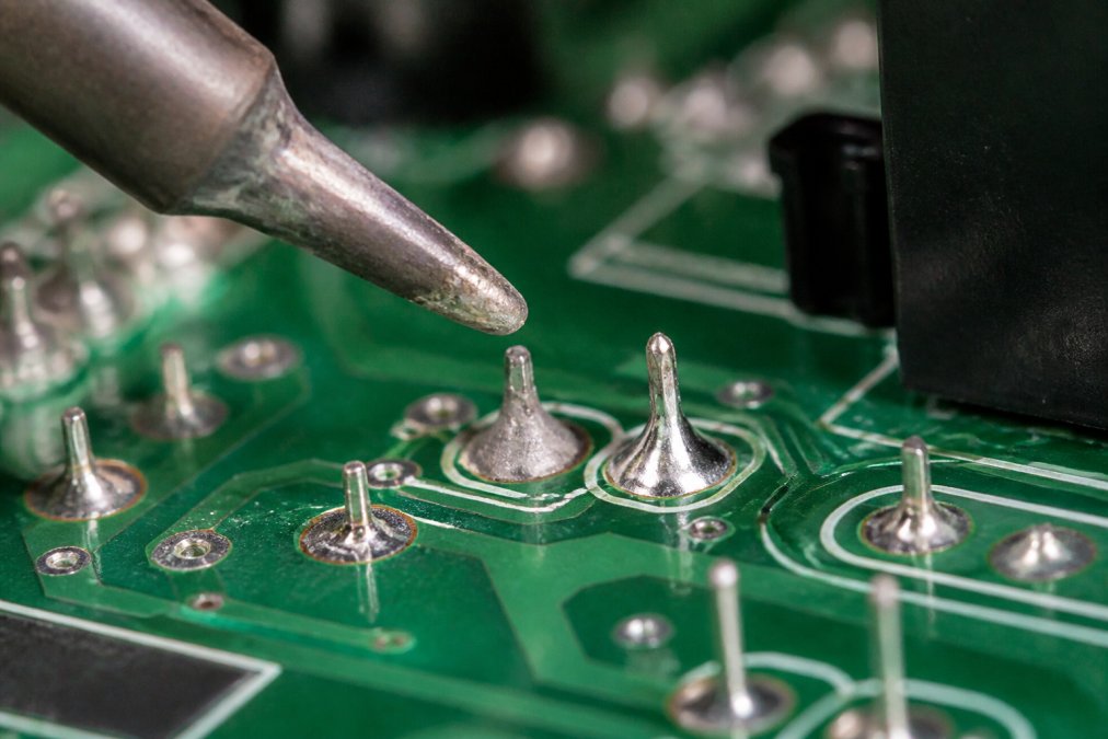 Macro photograph of a soldering iron tip hovering over a circuit board, showing one dull grainy solder joint next to a bright shiny joint under a bright work lamp