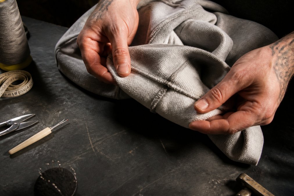 Close-up of hands turning a heavyweight cotton hoodie inside out on a dark workshop surface, examining the interior seam stitching and flat-felled seam detail under a focused overhead light