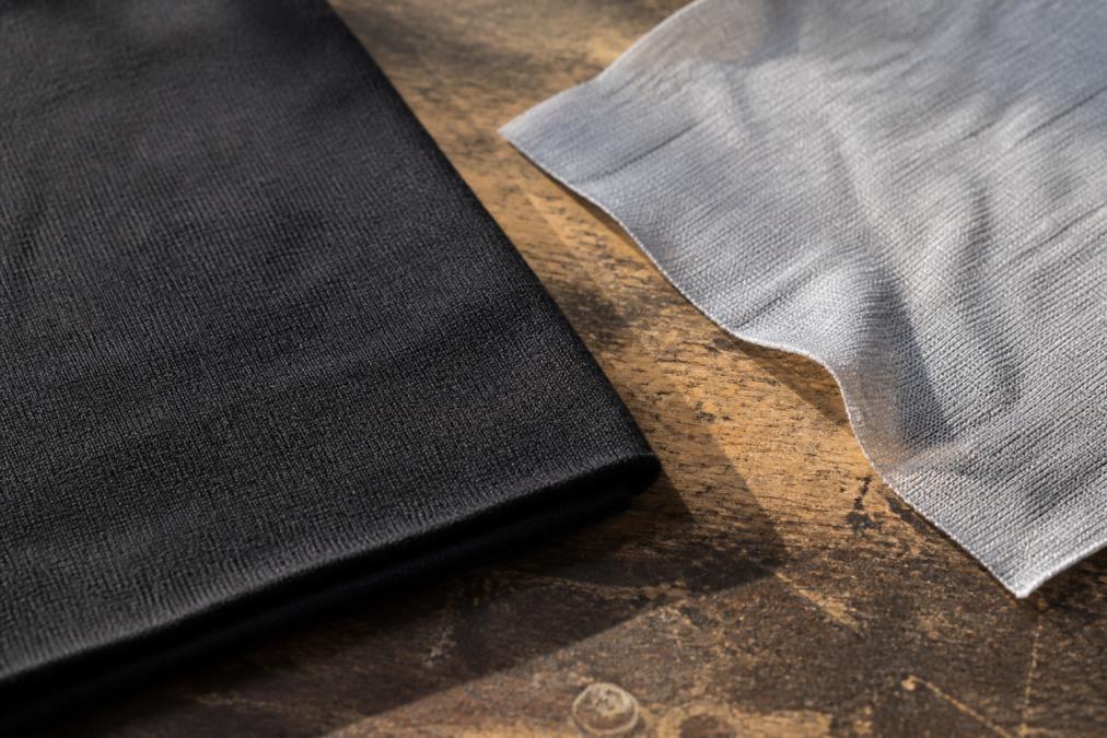 Close-up of two t-shirt fabric swatches laid flat on a worn wooden workbench, one visibly dense heavyweight ring-spun cotton with a tight weave, the other a thin synthetic blend showing slight transparency under natural window light