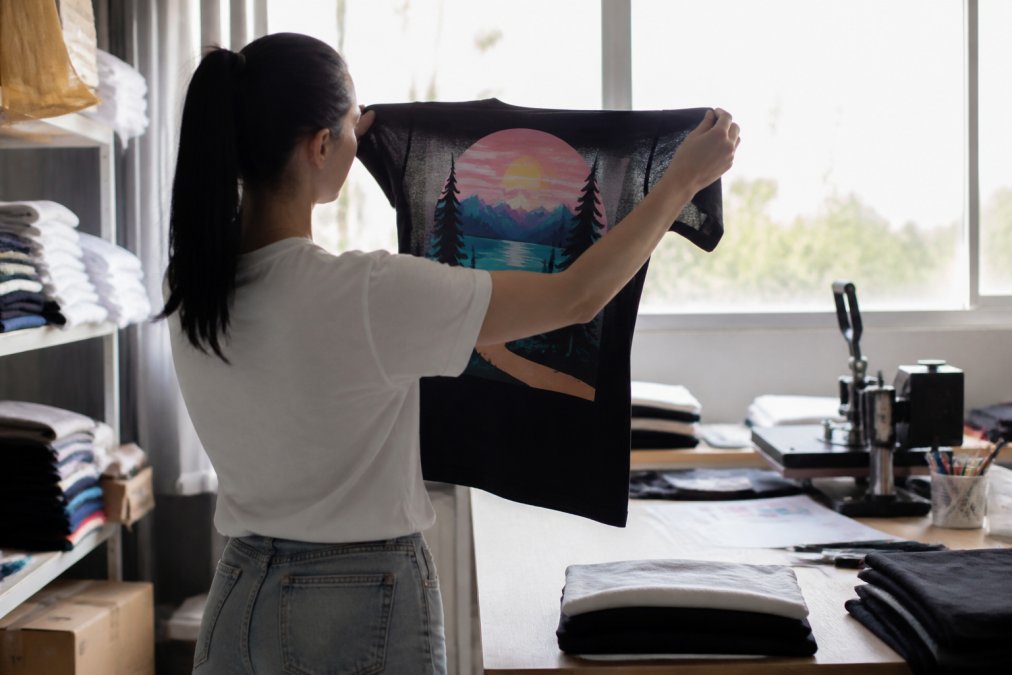 Hands holding a custom t-shirt up to natural light, closely examining the print edges for crispness and colour saturation, with the shirt partially backlit