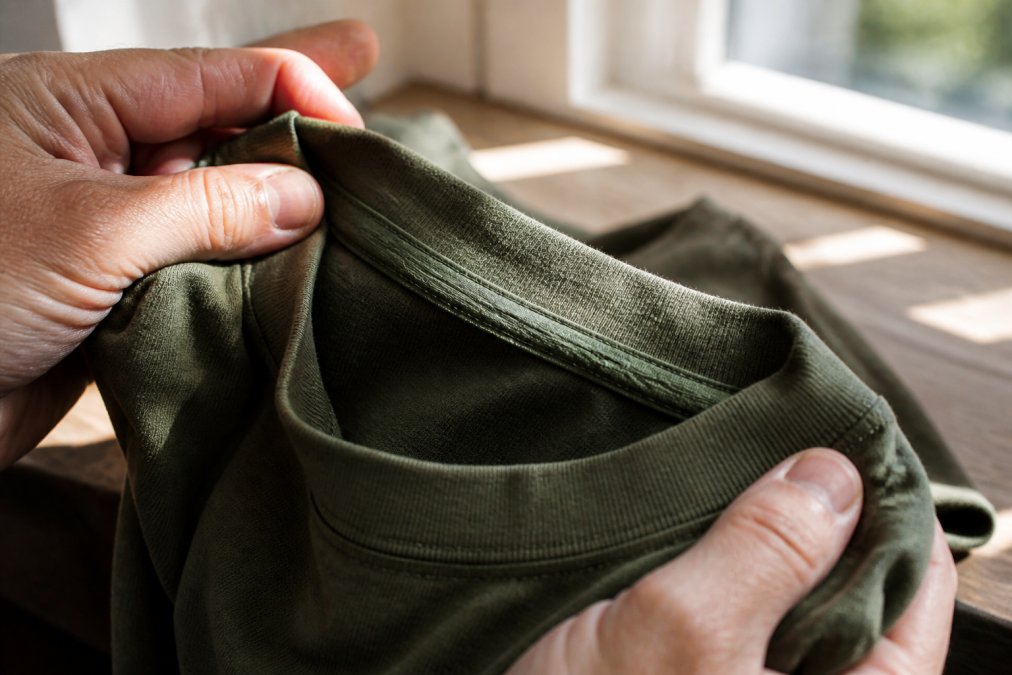 Close-up of hands inspecting the inside collar and shoulder seam of a folded heavyweight cotton t-shirt, showing the double-needle stitching and taped neckband under bright natural window light