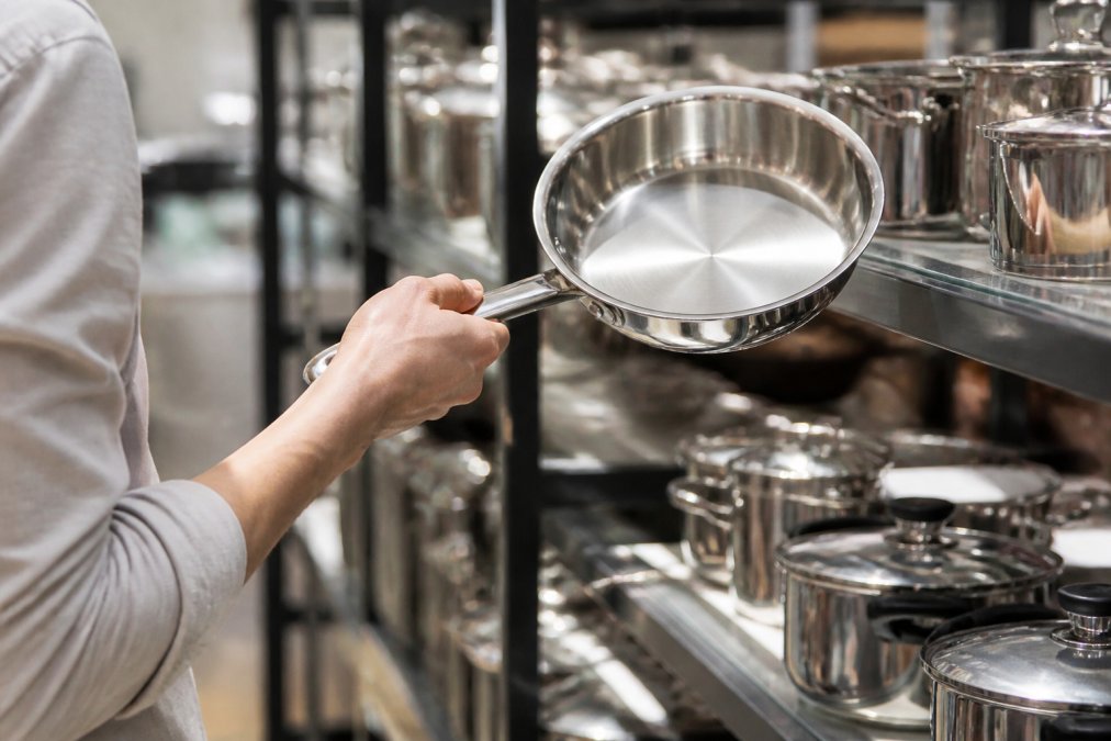 Close-up of a shopper's hand lifting a stainless steel pan to check its weight and balance under bright kitchen lighting
