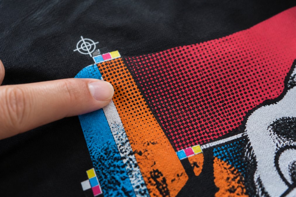 Close-up of a screen-printed t-shirt showing crisp, sharp halftone dots and registration marks with perfect color alignment under natural window lighting, with a finger pointing to the fine detail