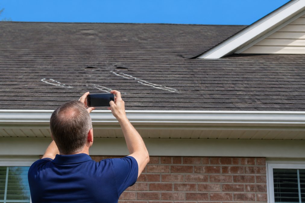 Homeowner photographing roof damage with a smartphone while standing safely on the ground, capturing visible shingle damage and dents in the frame with clear daylight