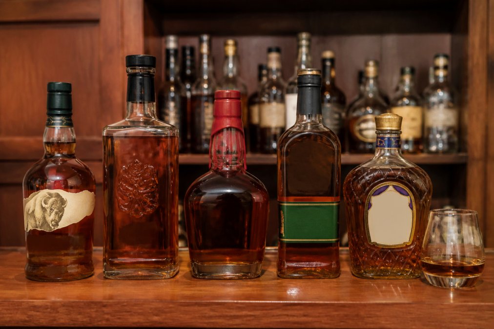 Five bourbon bottles arranged on a wooden shelf next to a glass of amber whiskey for a home bourbon collection.