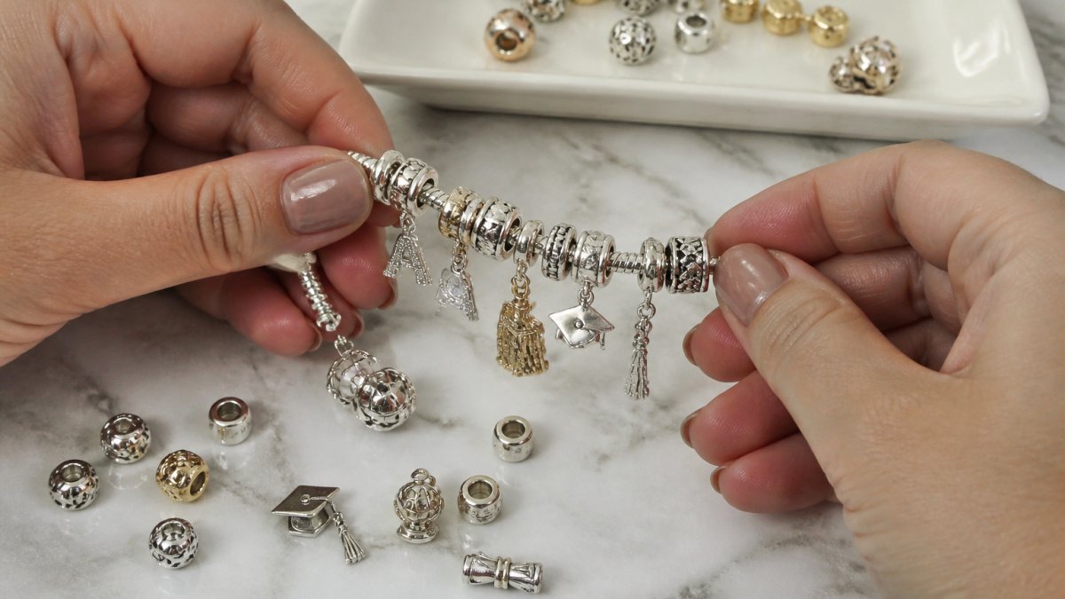Close-up of hands arranging sterling silver charms on a bracelet, showing various graduation-themed charms including a tassel charm and initial charm with white and gold plating visible