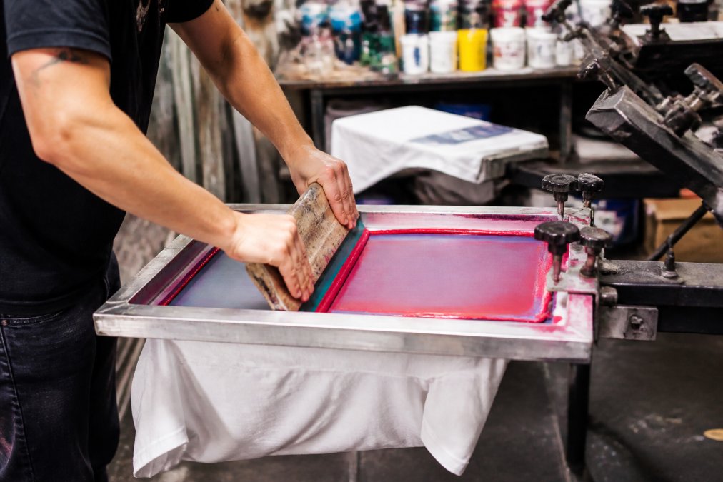 Close-up of hands pulling a squeegee across a screen on a manual press, with a white t-shirt underneath and bright plastisol ink visible on the mesh