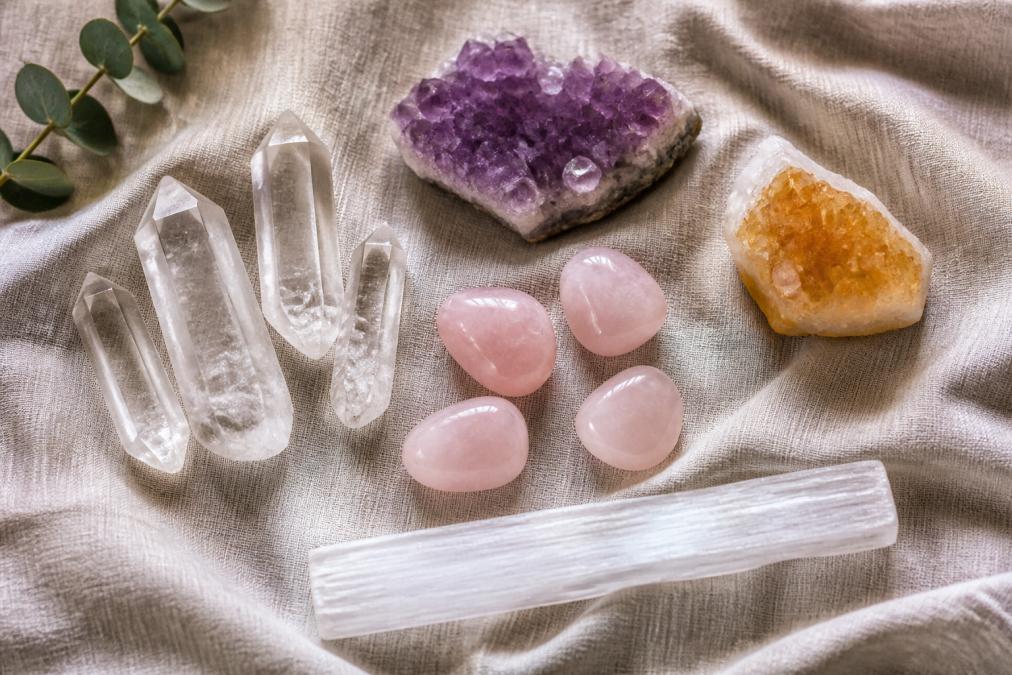 A flat-lay arrangement of beginner crystals including clear quartz points, amethyst clusters, rose quartz tumbles, citrine, and a selenite wand arranged on natural linen fabric under soft diffused window light