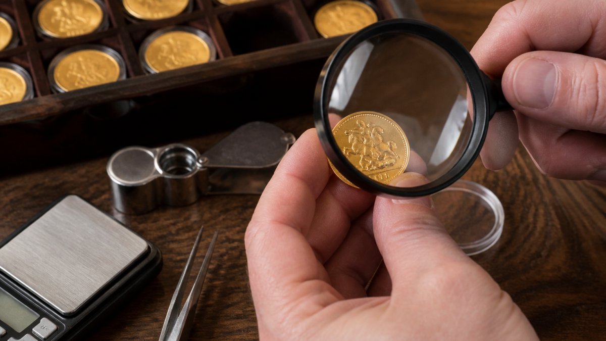 Hands using a magnifying glass to inspect the surface detail and mint marks of a gold sovereign coin under bright light.