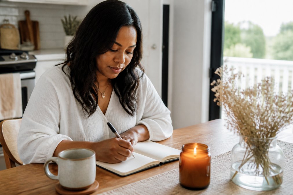 A woman in her thirties sitting at a sunlit kitchen table writing in a small notebook beside a warm mug and a lit amber jar candle