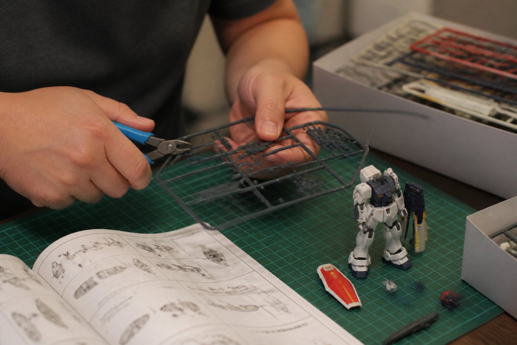 Close-up of a hobbyist's hands cutting a runner part from a sprue with nippers at a clean desk mat, with an instruction manual and a half-built Gundam model nearby