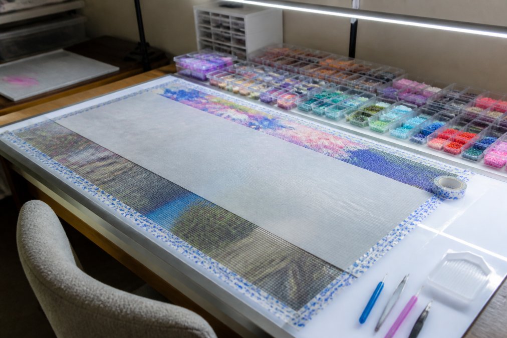 A crafting workspace with a large diamond painting canvas stretched on a light pad, organized trays of diamond drills in various colors, applicator tools arranged nearby, parchment paper, and washi tape securing the canvas edges under bright LED lighting