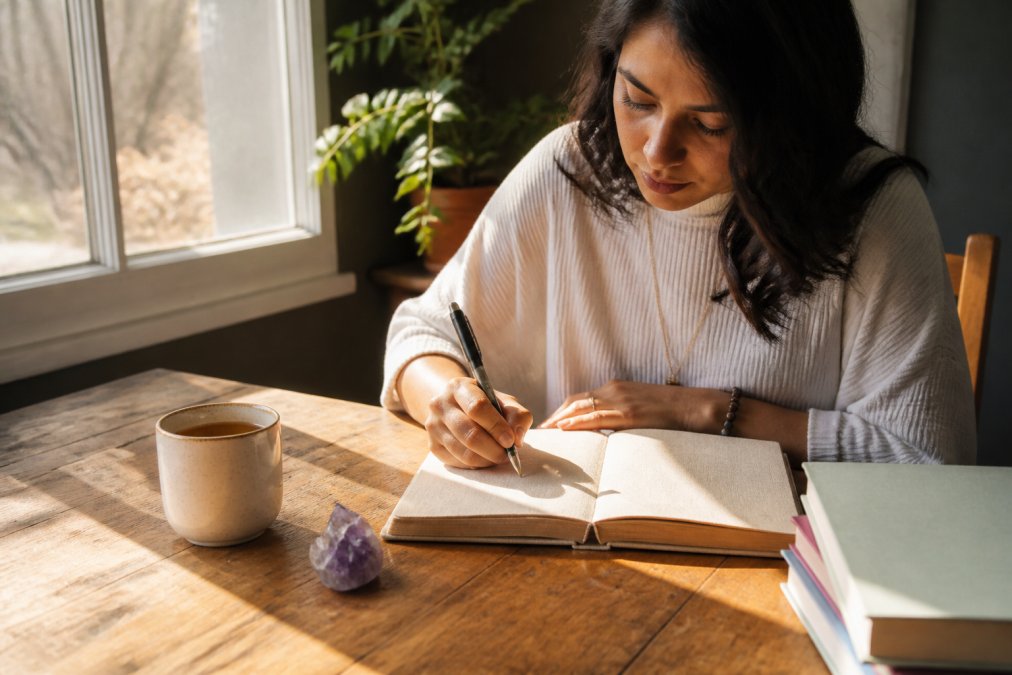 Woman writing in a journal with tea and a crystal as part of her daily spiritual practice in soft morning light.