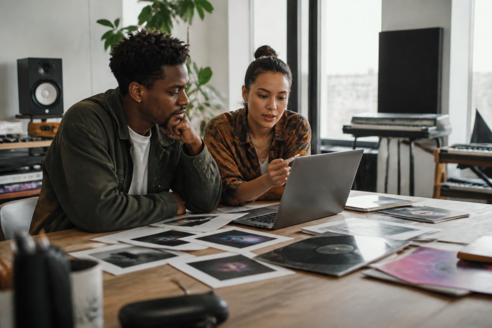 Music producer and artist reviewing album artwork on a laptop in a bright studio space, discussing visual branding elements for sublabel identity