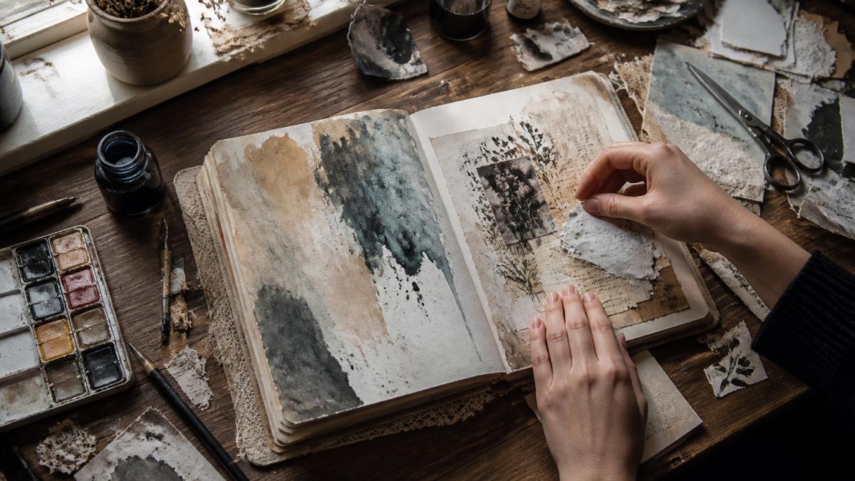 Overhead shot of hands creating an art journal page with watercolor washes, collage materials, and mixed media supplies spread across a wooden desk with natural window lighting